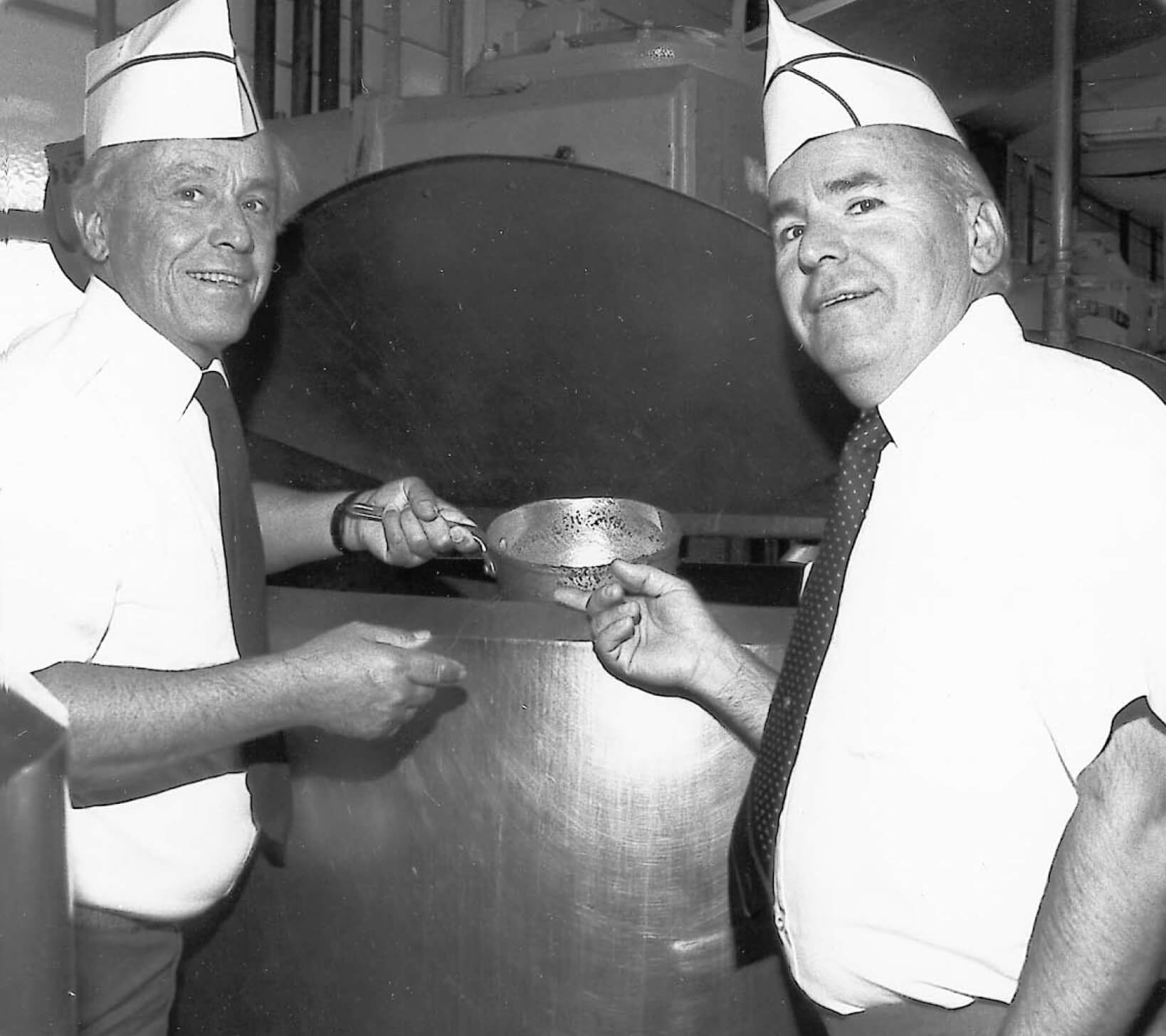 Two white men wearing short sleeved shirts, ties, and paper caps standing next to a chili kettle - photo is in black and white